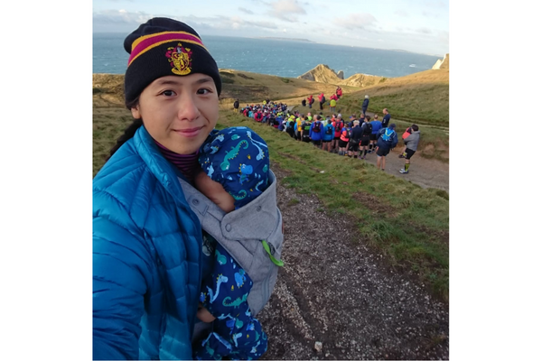 A lady in a beanie hat is standing holding her small baby in a dinosaur onesie in front of a group of runners on the Jurassic Coast